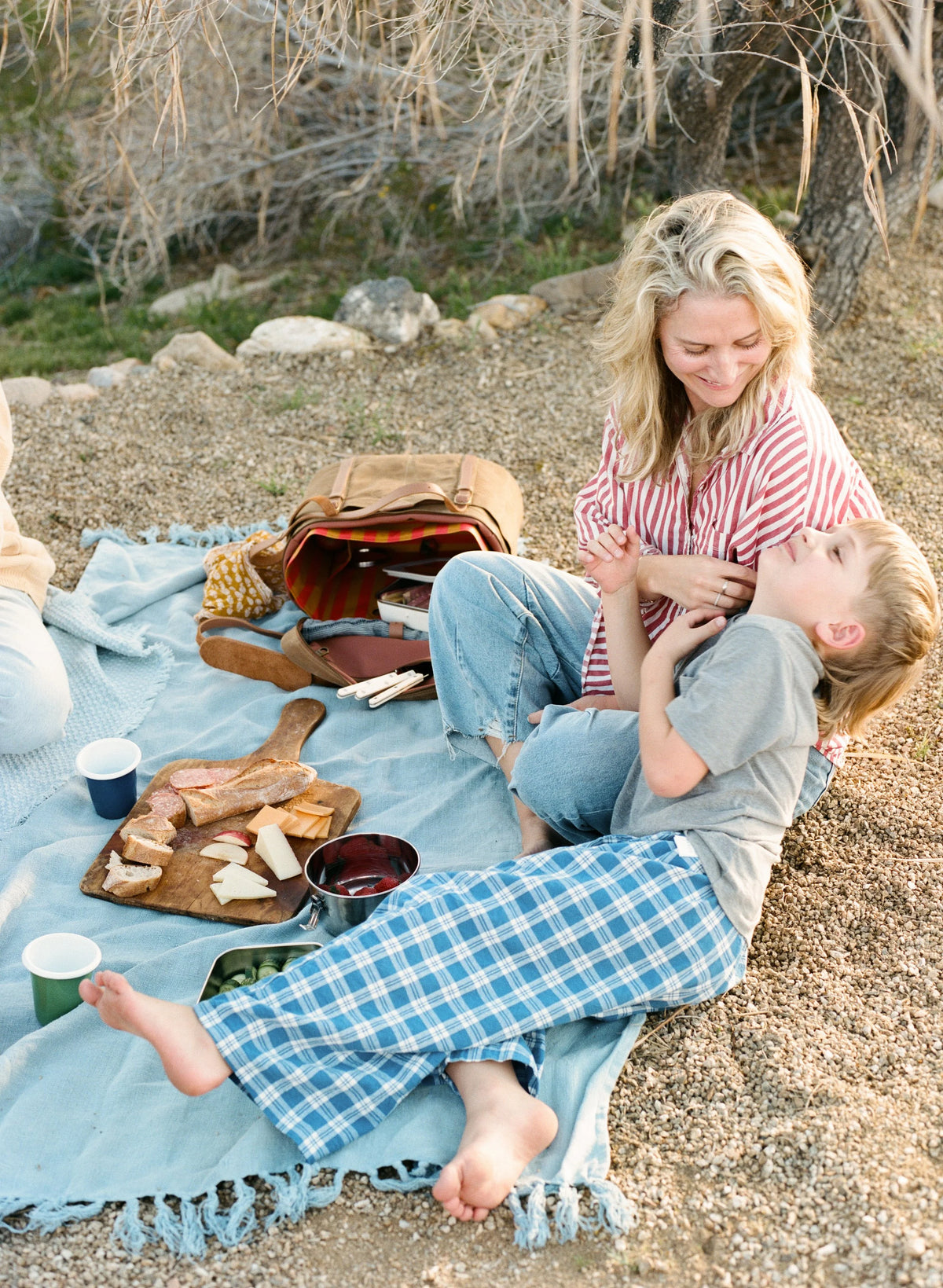 A mother and son relax on a blue picnic blanket outdoors, with a waxed canvas cooler bag, bread, and cups nearby. The woman wears a red-and-white striped shirt and blue jeans, and the boy wears a gray T-shirt and blue-and-white plaid PJ pants.